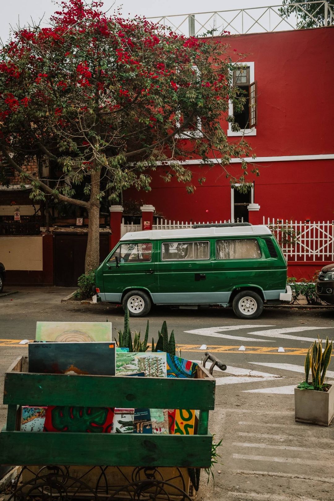 A green camper van parked in front of a red house in Barranco, with a bougainvillea in bloom and a cart of paintings for sale in the foreground