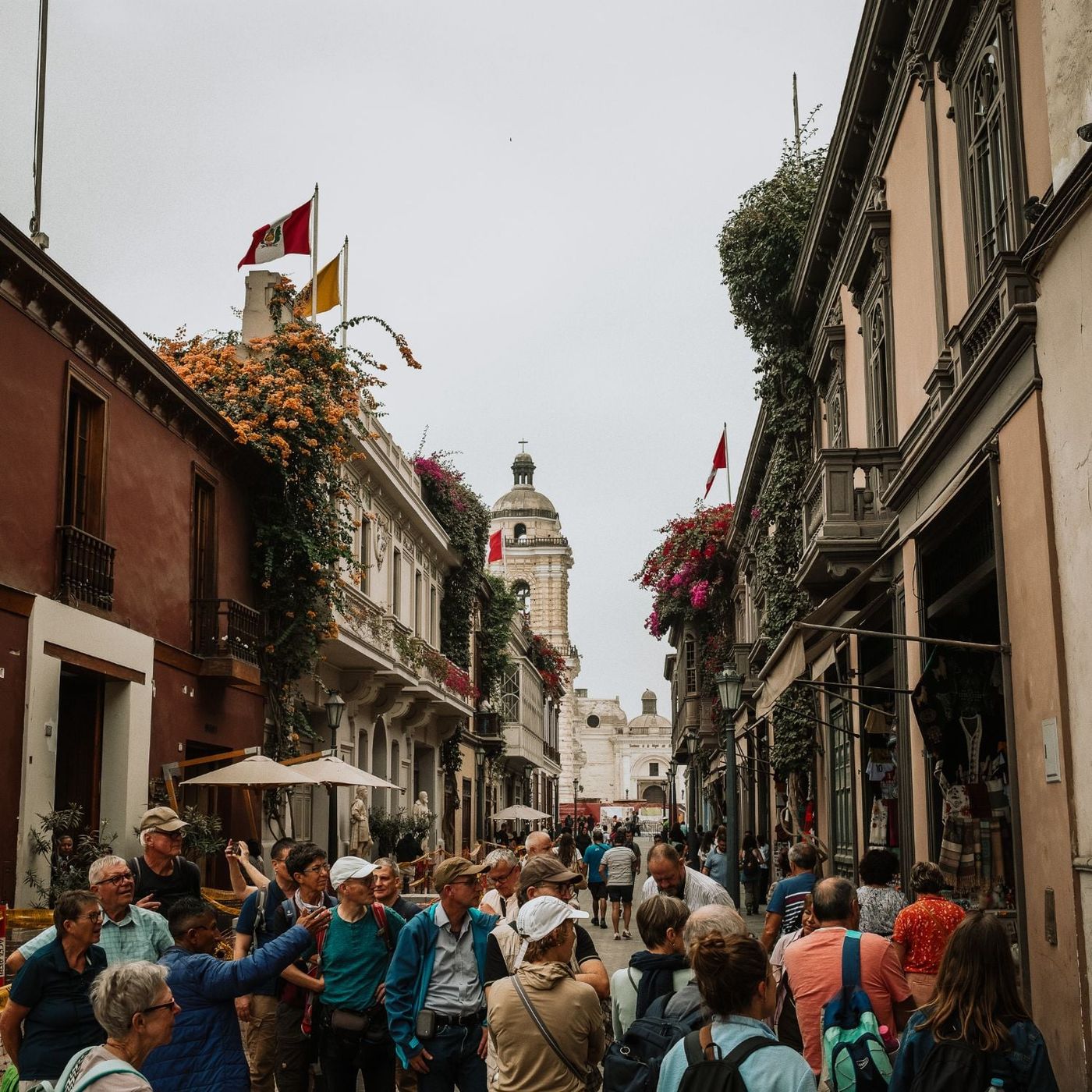 A busy street in central Lima with flowering bougainvillea on the balconies, Peruvian flags and a church bell tower at the far end, and visitors walking below