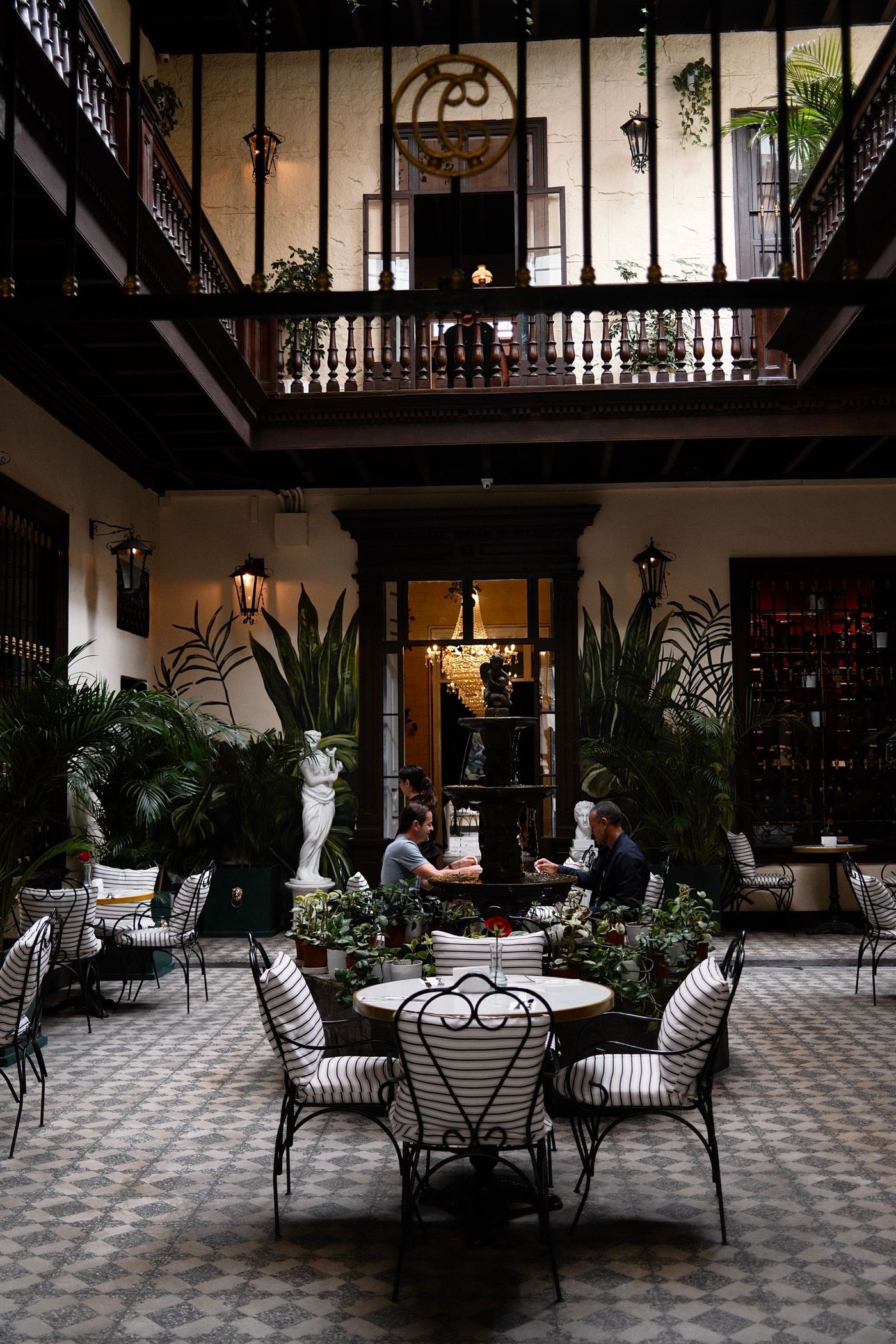 A quiet courtyard cafe with a stone fountain, palms and potted plants, striped iron chairs around a marble table and wooden balconies above