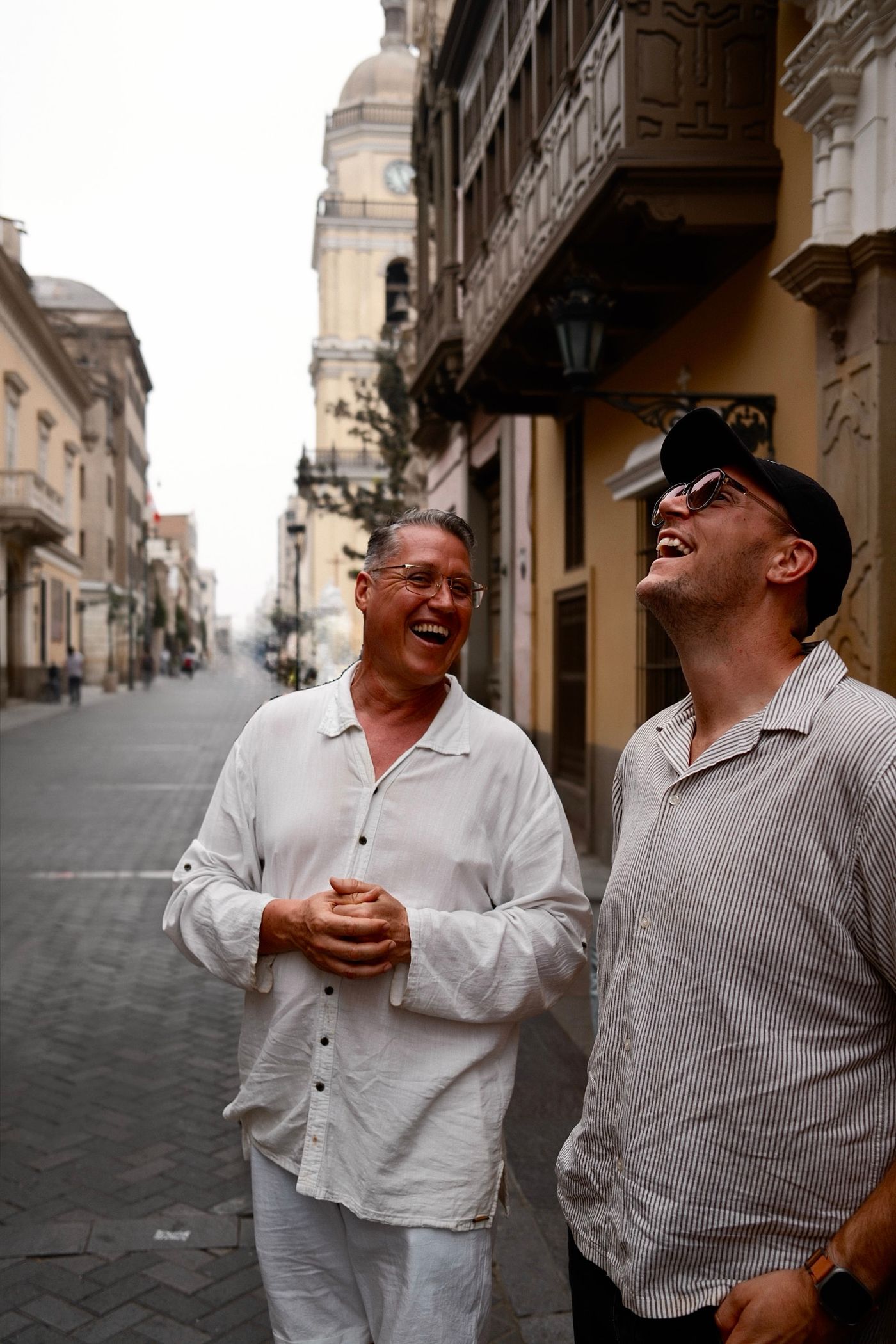 Manny laughing with a companion in a cap on a quiet cobbled street in Lima’s historic centre, with carved wooden balconies and a church tower with a clock behind them