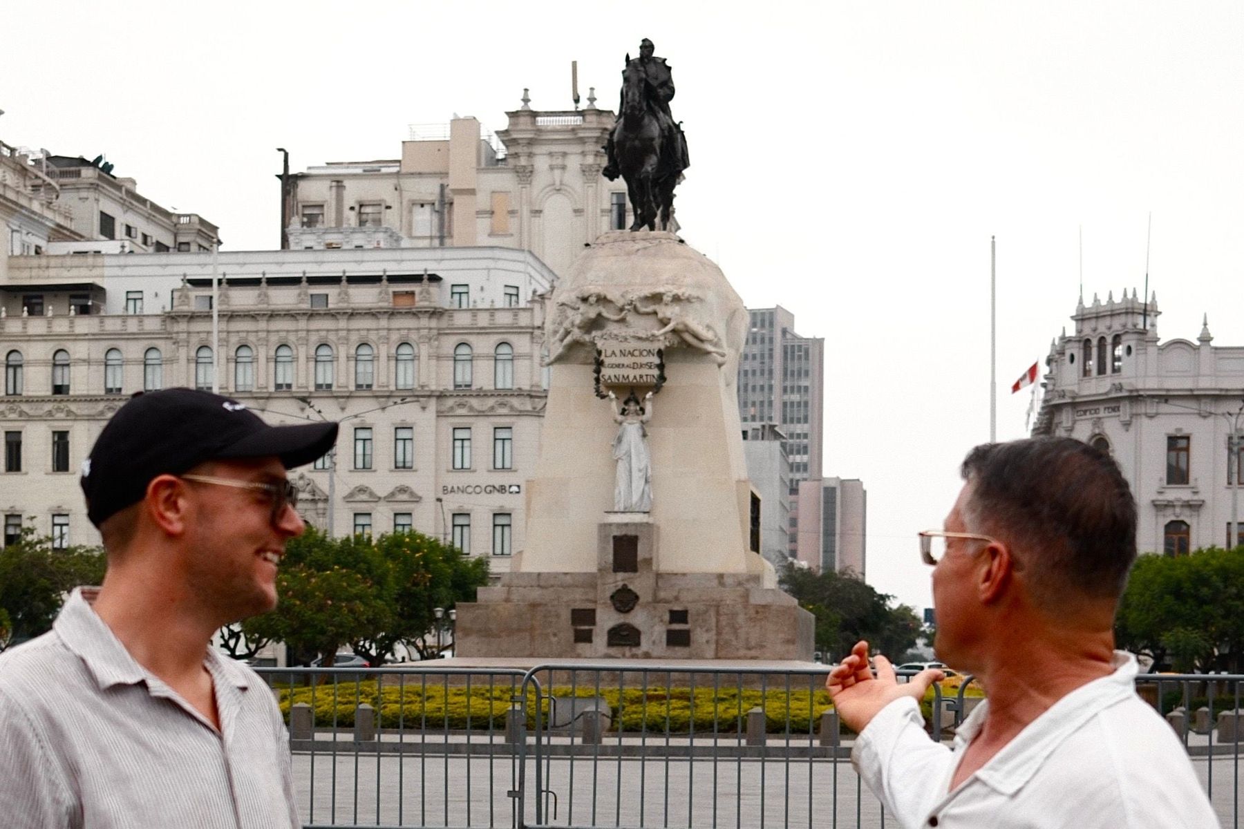 Manny in a white shirt pointing at the San Martín monument in Lima’s Plaza San Martín while a companion in a cap laughs beside him