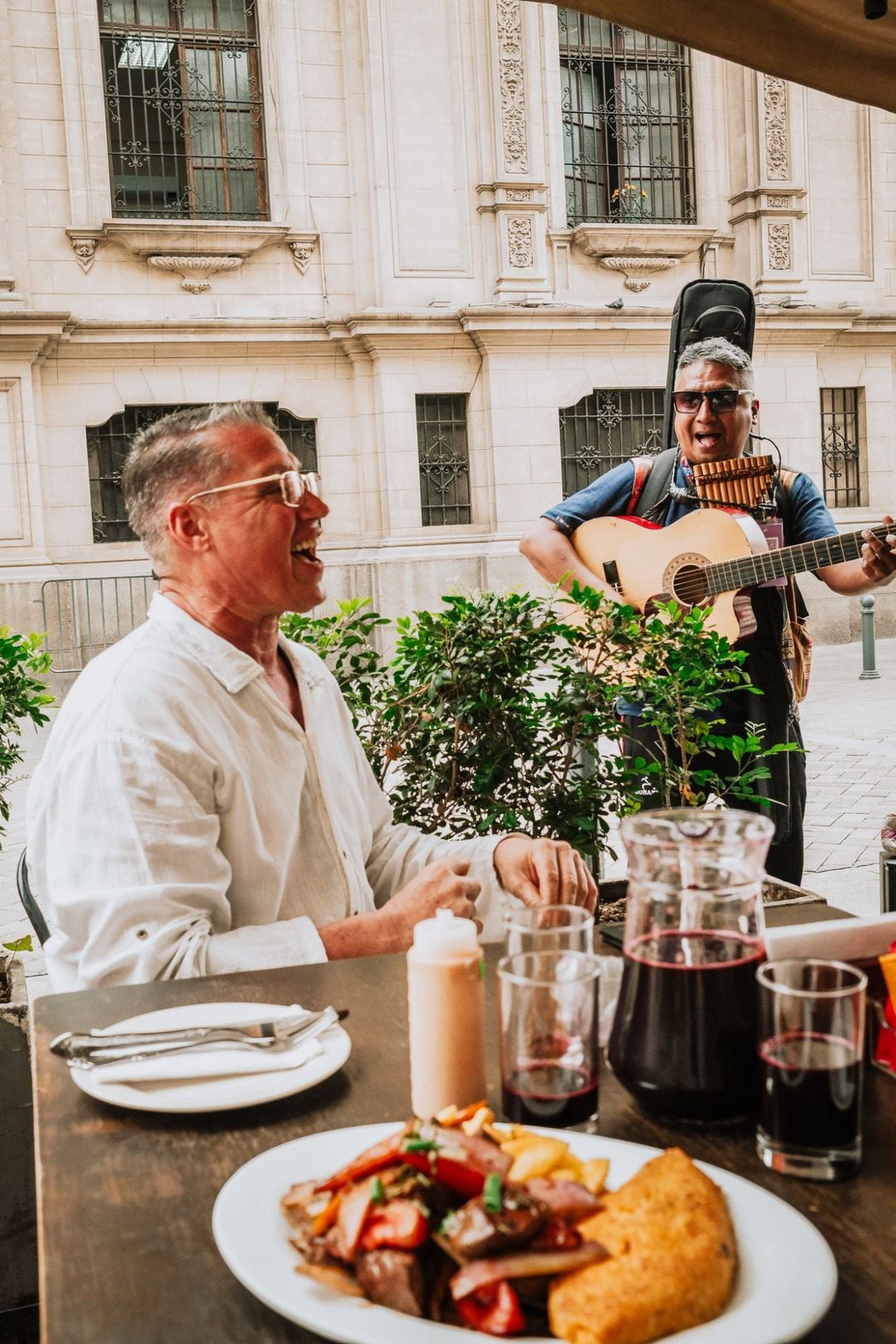Manny laughing at an outdoor table in Lima while a street musician with a guitar and pan pipes plays beside him, with a plate of lomo saltado in the foreground